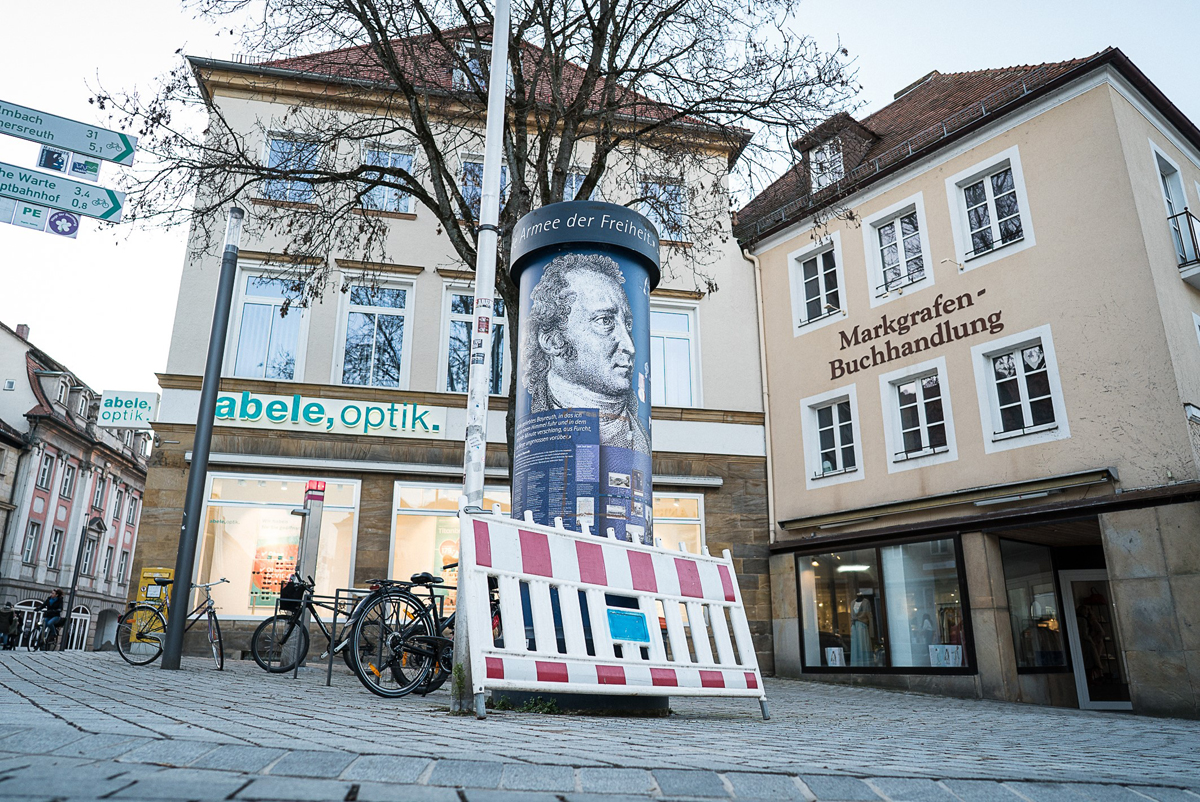 Litfaßsäule mit zwei Gebäuden im Hintergrund.