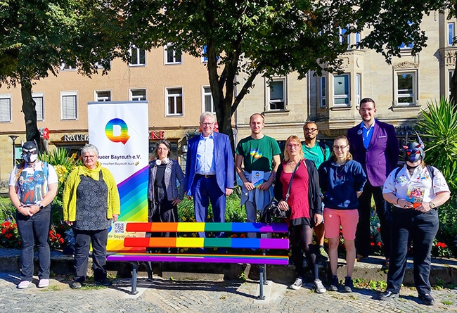 Mehrere Menschen stehen hinter der in Regenbogenfarben gestrichenen Bank