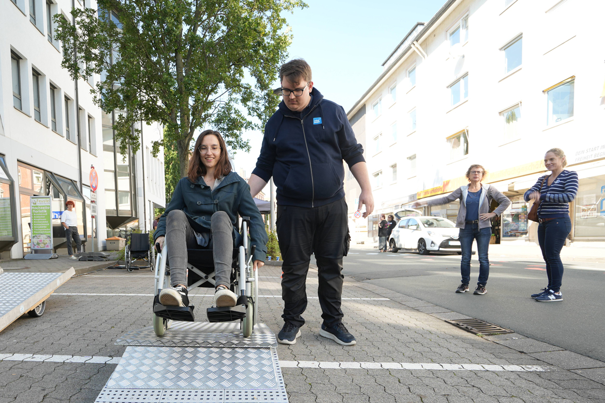 Eine Frau in einem Rollstuhl wird von einem Helfer beim Bewältigen einer Rampe unterstützt