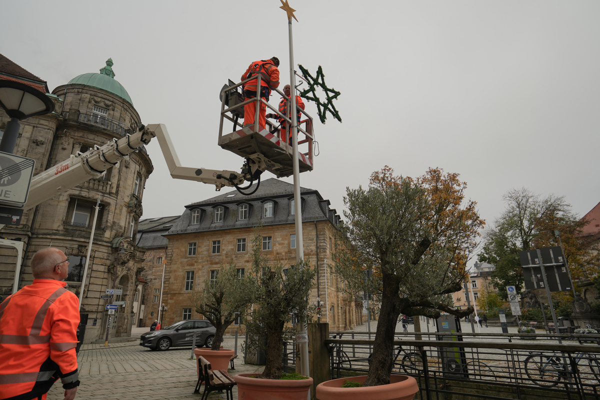 Zwei Mitarbeiter des Stadtbauhofs bringen mit Hilfe eines Krans einen Weihnachtsstern an einer Stange an. Ein weiterer Mitarbeiter sichert die Straße.