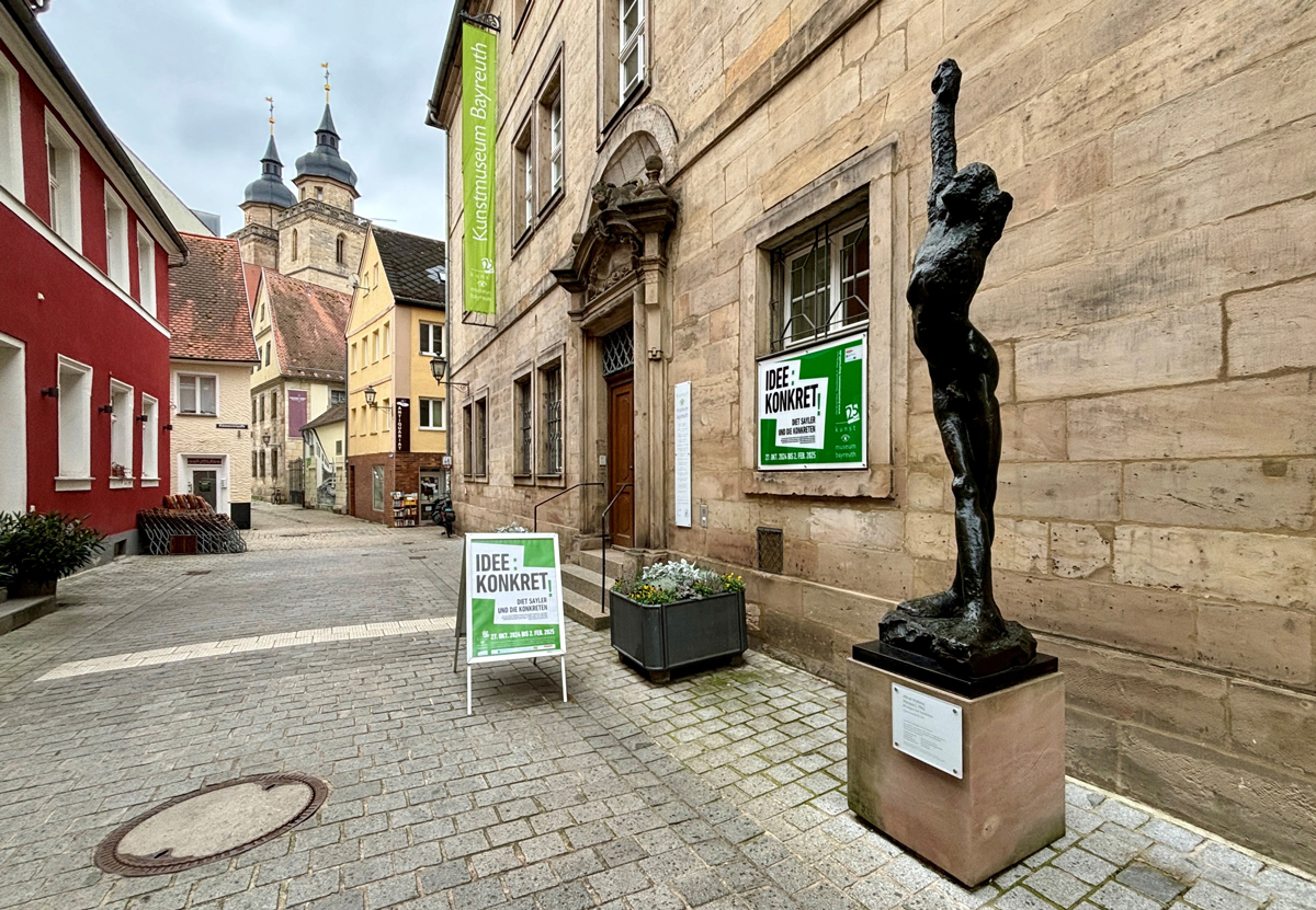 Der Eingang zum Kunstmuseum Bayreuth. Im Vordergrund ist eine Skulptur zu sehen, im Hintergrund die Türme der Stadtkirche.