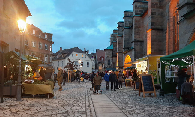 Das Bild zeigt einen Weihnachtsmarkt in einer historischen Innenstadt neben einer Kirche.