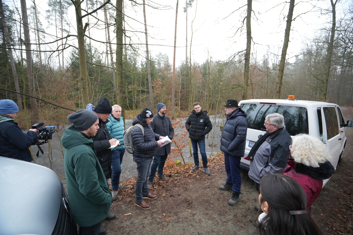 Eine Personengruppe steht im Wald und besichtigt einen zu rekultivierenden Teich.